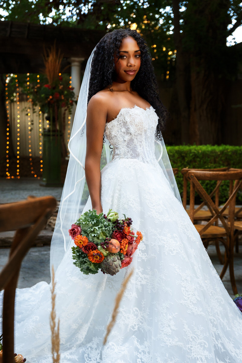 Full-length portrait of a smiling bride wearing a strapless lace gown from Just Girl Stuff, holding a vibrant bridal bouquet with greenery and artichokes by Lund Floral at Eldridge Manor in Ogden, Utah.