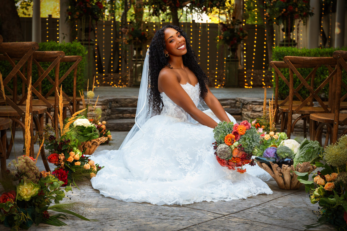 Seated portrait of a smiling bride holding a vibrant bridal bouquet with greenery and artichokes by Lund Floral, wearing a strapless lace gown from Just Girl Stuff at Eldridge Manor in Ogden, Utah.
