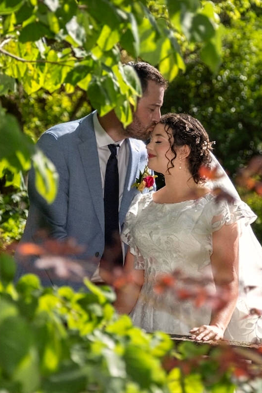 Groom kissing the bride's forehead gently outdoors framed by green foliage and natural light.