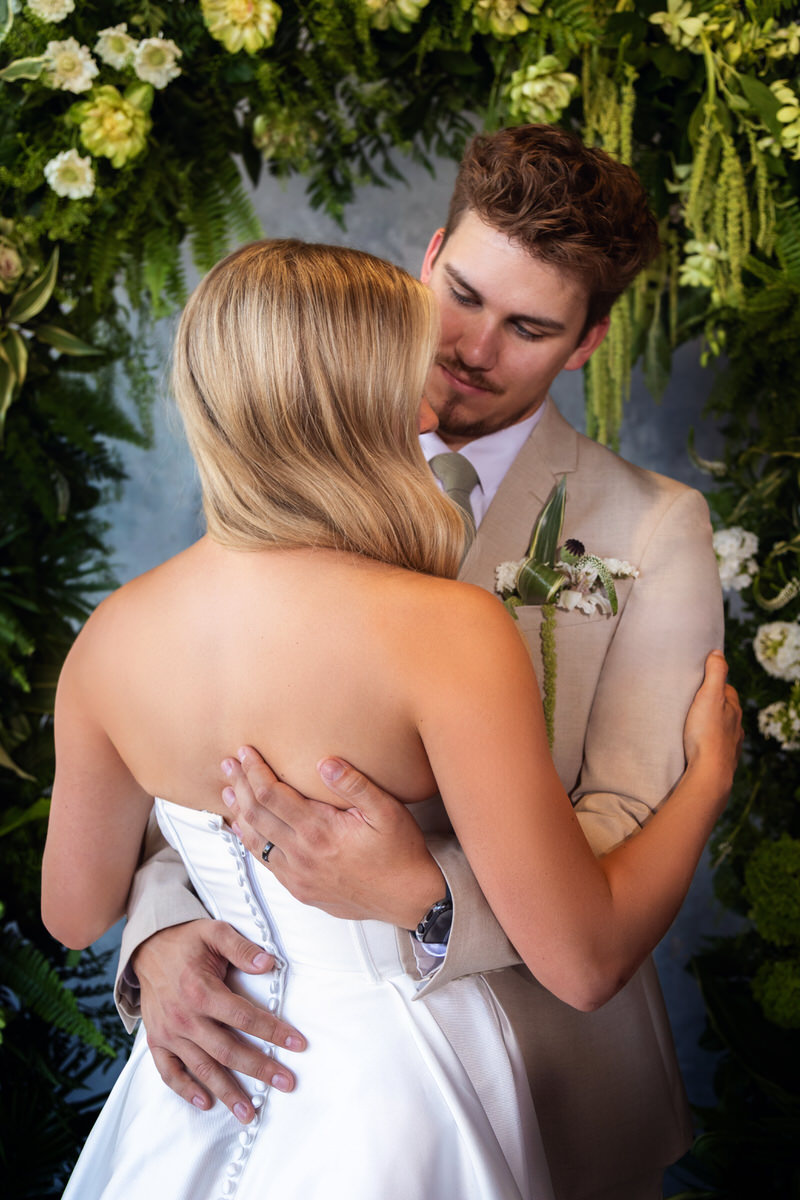 Groom in a tan suit holding and embracing a bride in a white strapless wedding gown, framed by an archway of lush green foliage and white flowers.