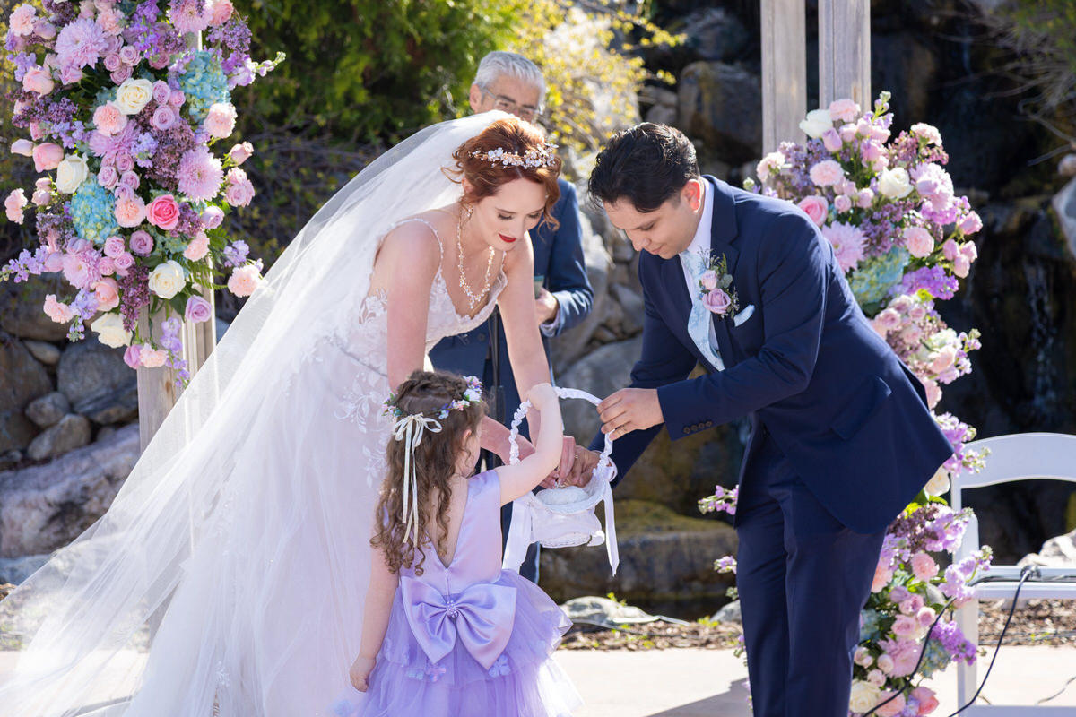 Bride and groom taking rings from a basket held by a young flower girl during an outdoor wedding ceremony flanked by floral arrangements.