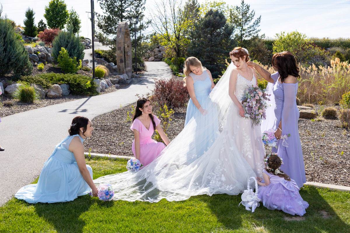 Bride in a white gown and veil getting ready outdoors, surrounded by bridesmaids in pastel dresses and a flower girl on a sunny garden pathway.