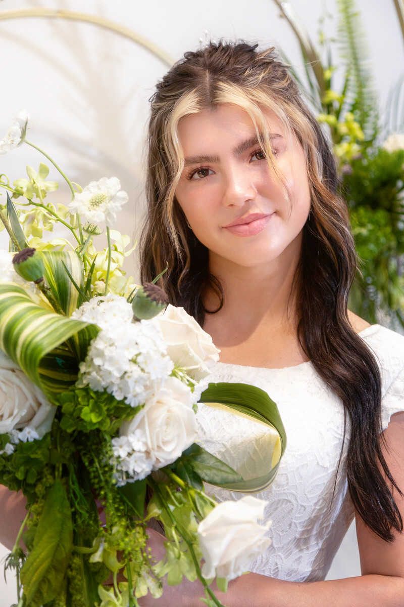 Groom in a tan suit holding and embracing a bride in a white strapless wedding gown, framed by an archway of lush green foliage and white flowers.