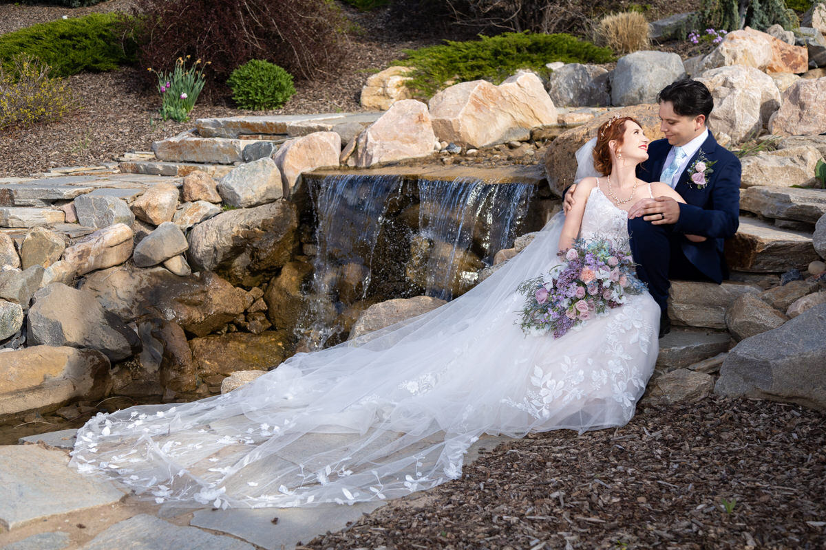 Bride and groom sitting on stone steps beside a cascading waterfall, with the bride holding a pastel floral bouquet and wearing a long veil spread out on the ground.