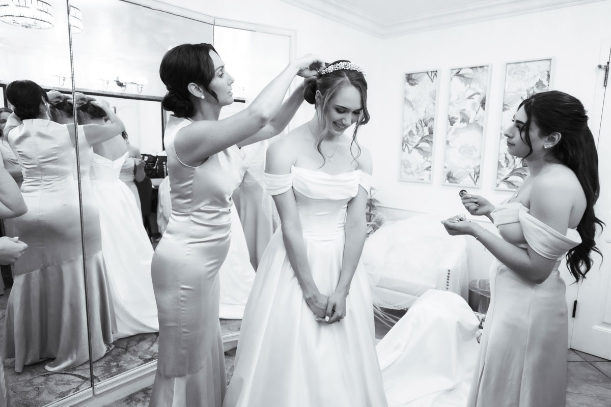 A woman in a wedding dress having her headpiece adjusted by a bridesmaid, with another bridesmaid holding a small object nearby, in a black and white photo.