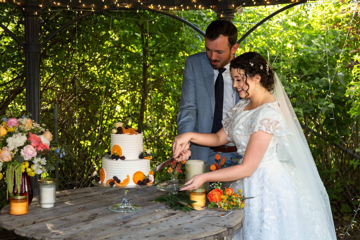 Bride and groom cutting a tiered wedding cake decorated with fresh oranges and berries underneath a gazebo strung with lights.