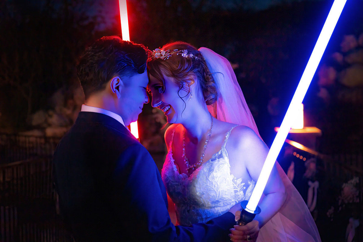 Bride and groom facing each other smiling at night, illuminated by glowing red and blue light saber props.