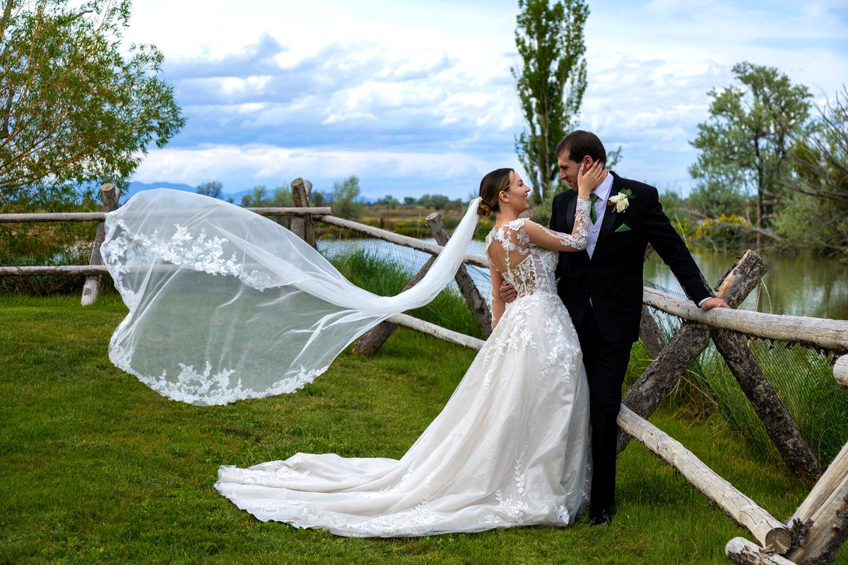 Bride and groom posing outdoors by a rustic wooden fence near water, with the bride's long lace-trimmed veil blowing dramatically in the wind.