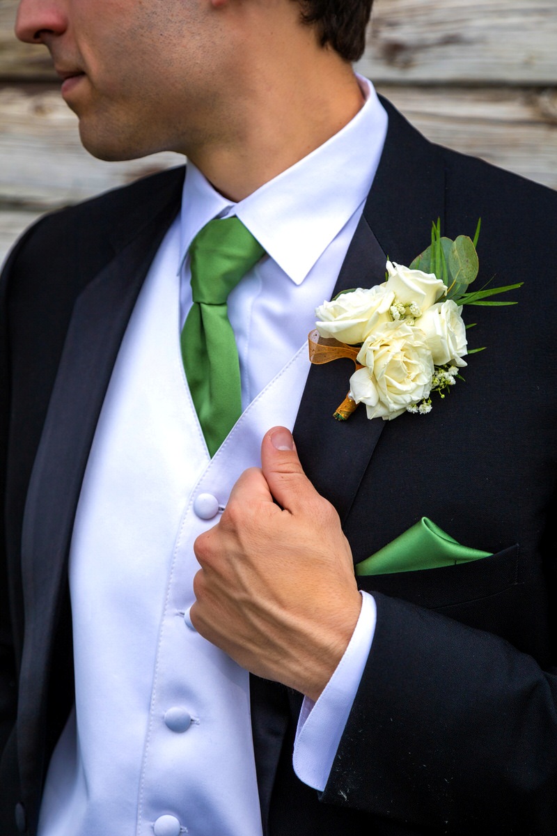 Close-up of a groom wearing a black tuxedo, white vest, green tie, pocket square, and a white rose boutonnière.