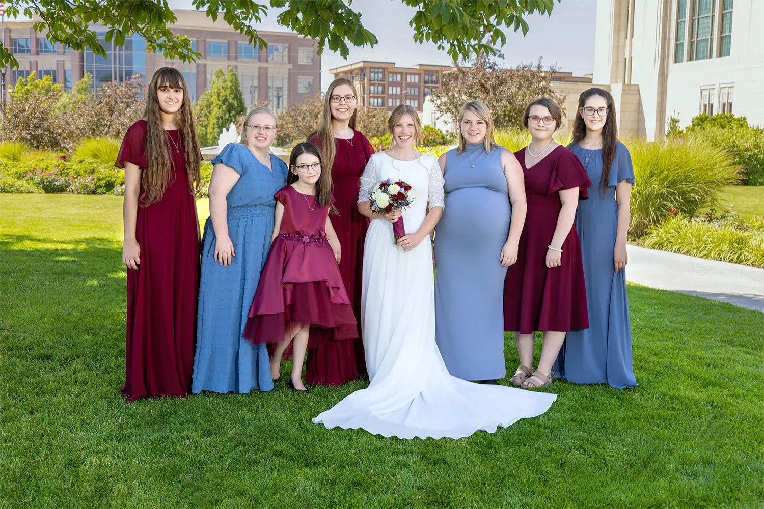 Bride in a white gown holding a bouquet standing with six bridesmaids and a younger attendant in burgundy and blue dresses outdoors on a lawn
