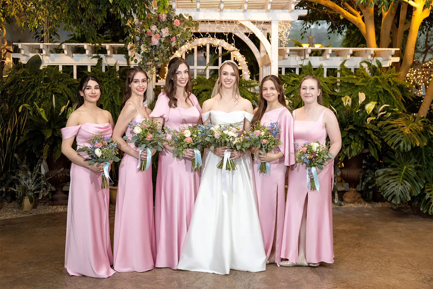 Bride in an off-the-shoulder white gown holding a bouquet standing with five bridesmaids in matching pink dresses in front of a white wooden trellis and greenery