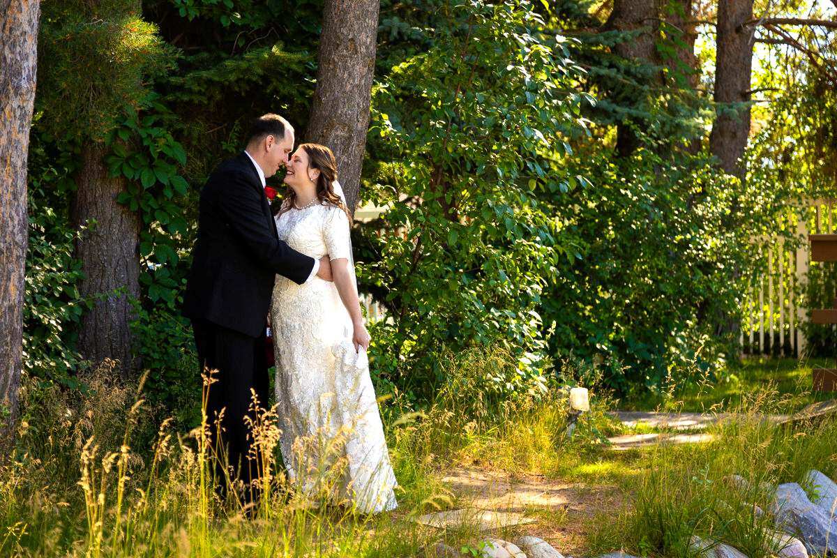 Groom in a black suit embracing and smiling at the bride in a white lace wedding gown outdoors near trees and tall grass.
