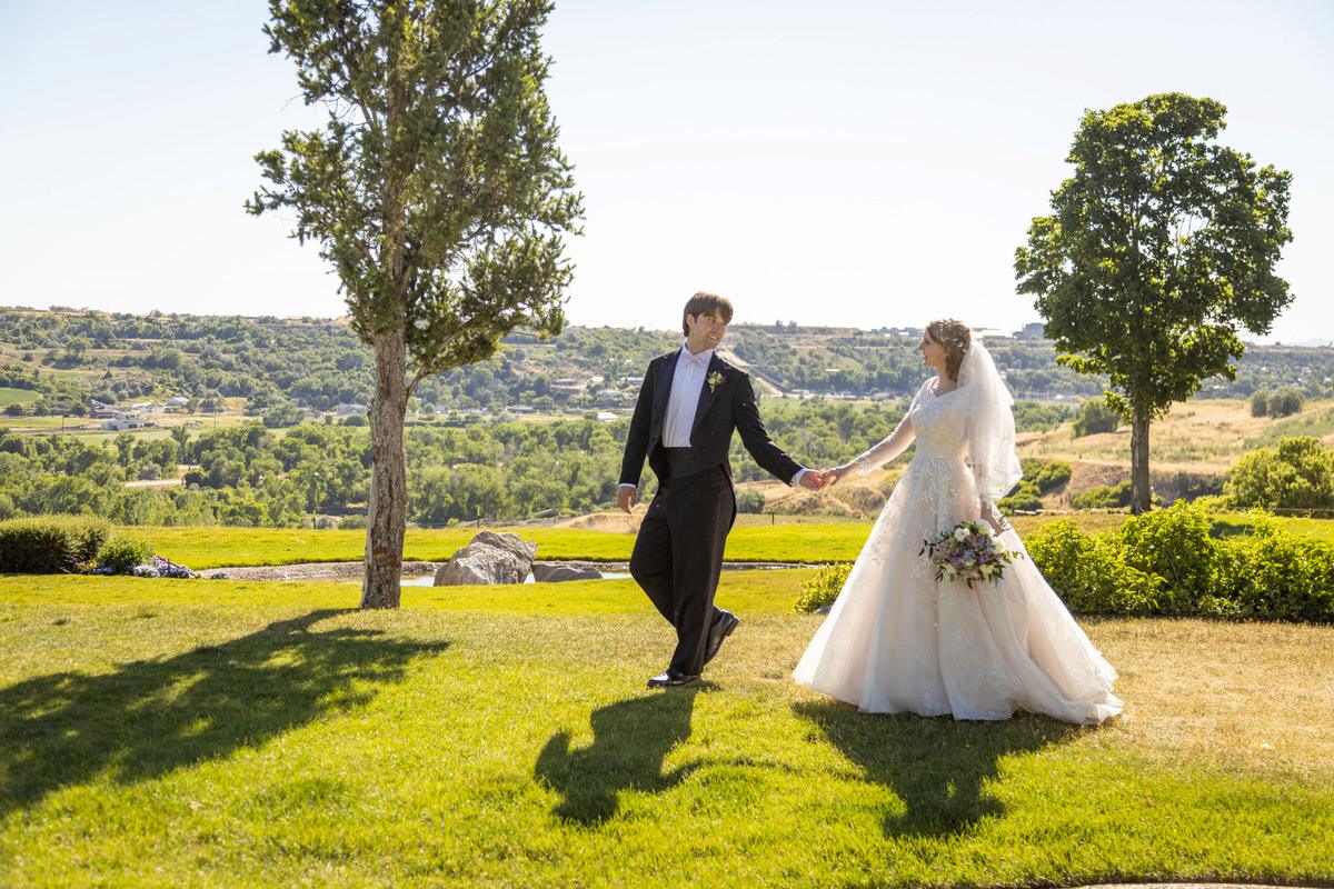 Bride and groom walking hand-in-hand down an outdoor pathway surrounded by lush greenery and warm sunlight.
