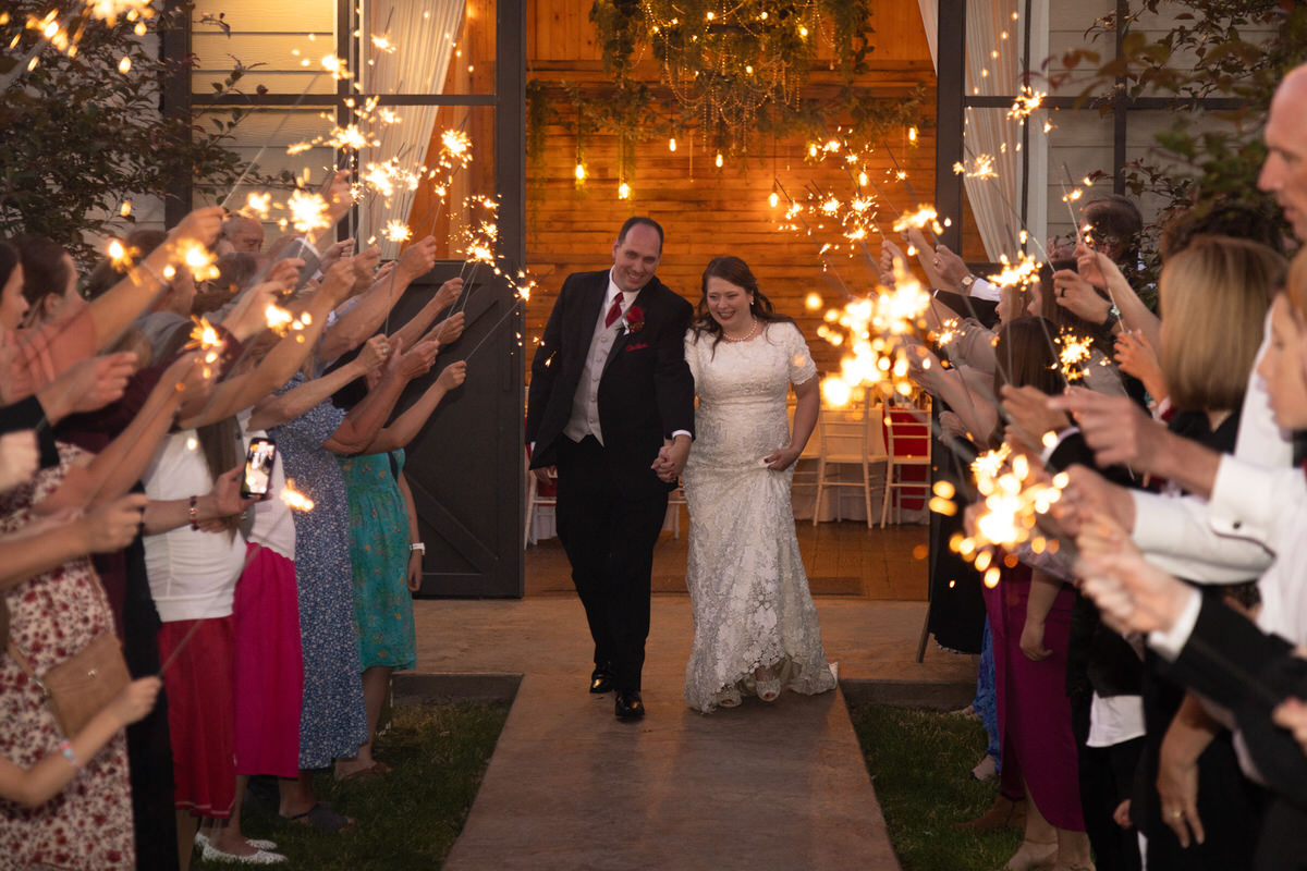 Bride and groom walking hand-in-hand down a pathway exiting through a tunnel of guests holding lit sparklers outside a venue with a wooden barn door backdrop.