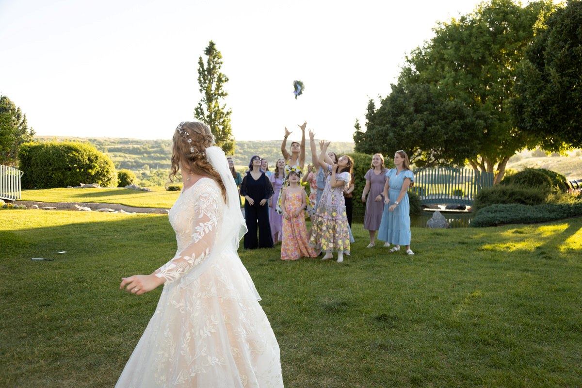 Bride tossing her bouquet outdoors in a sunlit garden surrounded by cheering wedding guests.