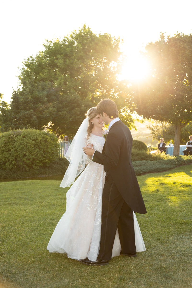 Bride and groom dancing outdoors in a sunlit garden with warm sunlight filtering through the trees behind them.