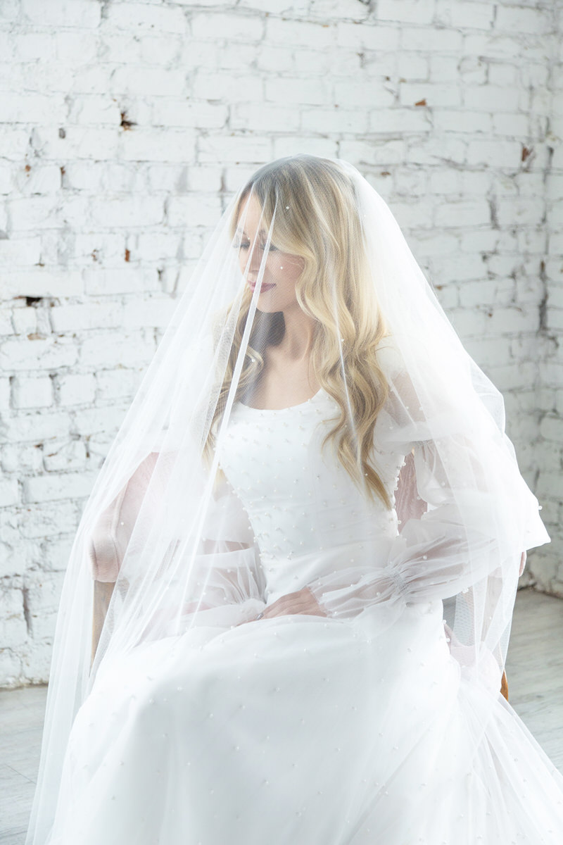 Bride wearing a white gown and a sheer veil with pearl accents, sitting in front of a white brick wall.