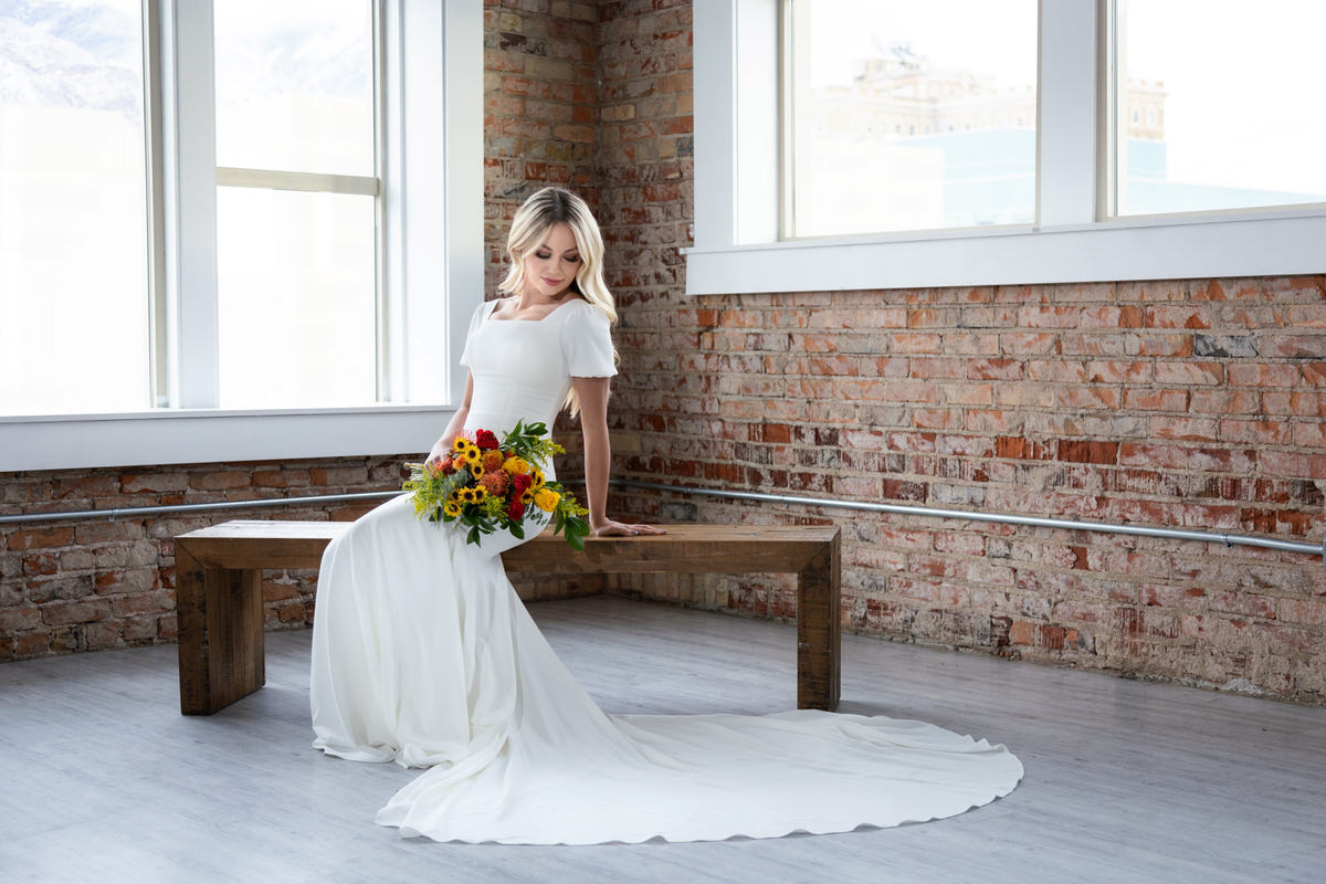 Bride wearing a white gown holding a vibrant bouquet of sunflowers, sitting on a wooden bench in front of an exposed brick wall and large windows.