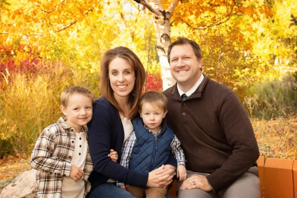 A family of four smiling together outdoors surrounded by autumn fall foliage during a portrait session at Masterpiece Images in Ogden, Utah.