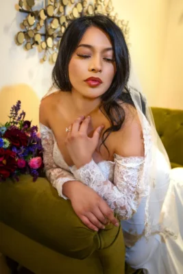 Bride posing indoors on a stylized lounge couch with a floral bouquet, resting her hand near her chest.