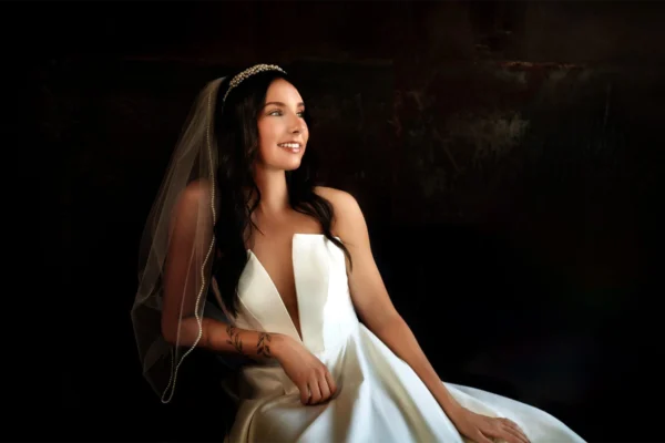 Bride in a wedding dress with a veil standing against a dark, moody background.