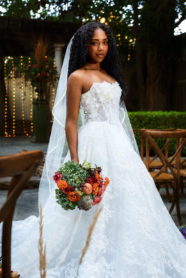 Full-length portrait of a smiling bride wearing a strapless lace gown from Just Girl Stuff, holding a vibrant bridal bouquet with greenery and artichokes by Lund Floral at Eldridge Manor in Ogden, Utah.