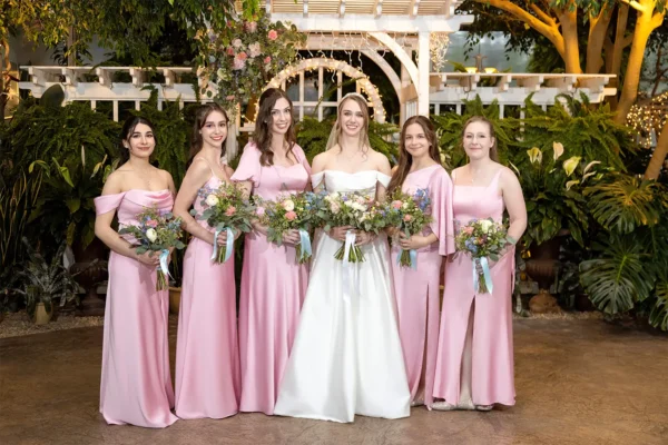 Bride in an off-the-shoulder white gown holding a bouquet standing with five bridesmaids in matching pink dresses in front of a white wooden trellis and greenery