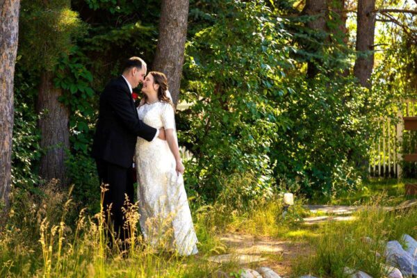 Groom in a black suit embracing and smiling at the bride in a white lace wedding gown outdoors near trees and tall grass.
